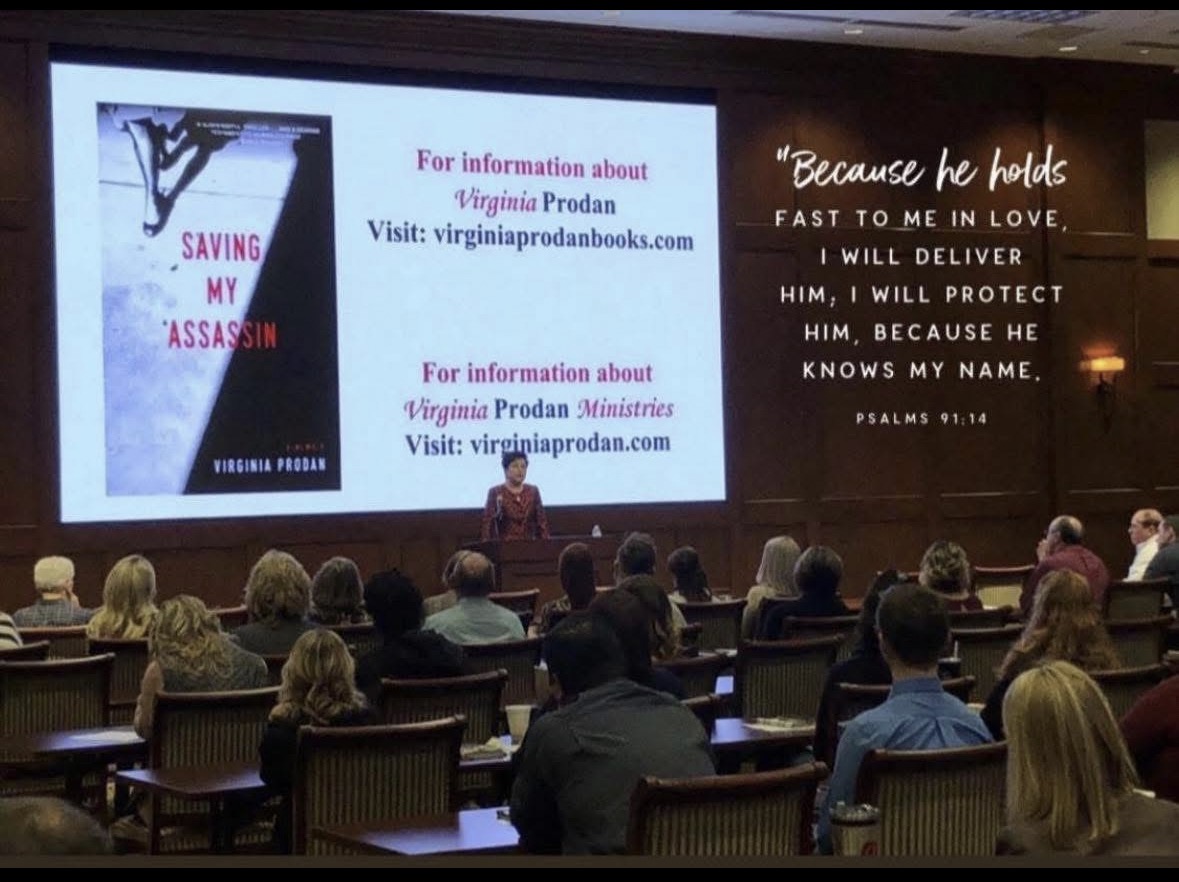 Virginia Prodan addressing an audience at Hobby Lobby, with Saving My Assassin displayed on the presentation screen.